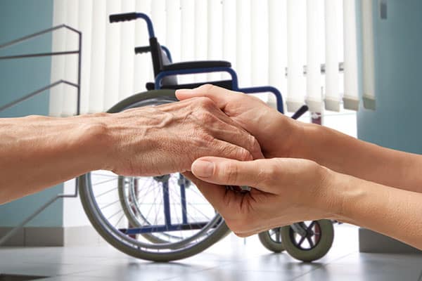 אישה מחזיקה ידיים של אישה מבוגרת וכסא גלגלים ברקע